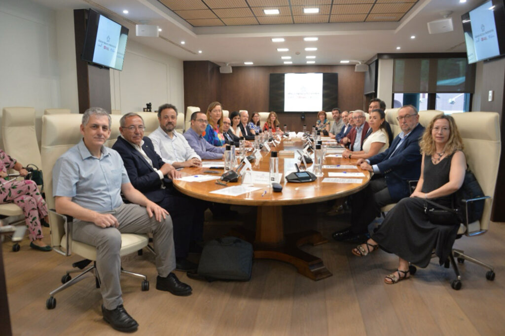 Un grupo de personas se sienta alrededor de una gran mesa de conferencias en una moderna sala de reuniones, mirando a la cámara. En cada extremo de la sala se ven pantallas de presentación. El ambiente parece formal y de negocios. Alianza Apoyo y Autonomía