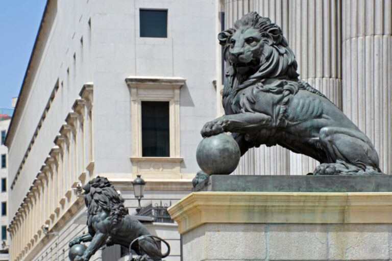 Dos estatuas de león de bronce descansan sobre pedestales de piedra en el exterior de un gran edificio con altas columnas. Un león apoya una pata en una esfera. La arquitectura es neoclásica, con grandes columnas estriadas y detalles ornamentales.