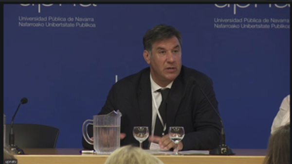 Un hombre trajeado sentado en una mesa con un micrófono, una jarra de cristal y dos vasos, hablando en una conferencia. El fondo muestra el logotipo de la Universidad Pública de Navarra sobre un fondo azul. Congreso Internacional sobre la Ley 8/2021