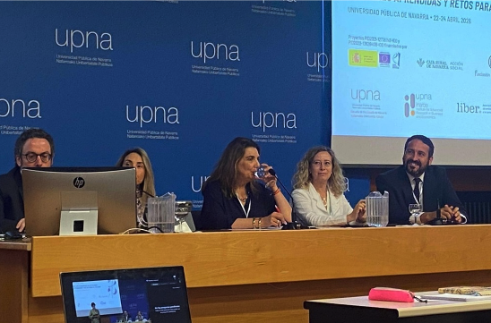 Cinco personas se sientan en una mesa frente a un telón de fondo azul con logotipos de "upna". Una mujer bebe agua mientras los demás sonríen, mientras se muestra una presentación en una pantalla a la derecha. Congreso Internacional sobre la Ley 8/2021