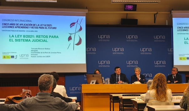 Cuatro ponentes se sientan en una mesa redonda frente al público durante una conferencia. Detrás de ellos, una gran pantalla muestra información sobre la Ley 8/2021 y los retos del sistema judicial. El acto se celebra en la UPNA. Congreso Internacional sobre la Ley 8/2021