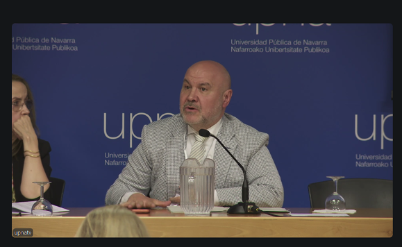 Un hombre calvo con un traje claro habla por un micrófono en una mesa de conferencias. El fondo muestra el logotipo de la Universidad Pública de Navarra sobre una pared azul, con una jarra de cristal y dos vasos sobre la mesa. Congreso Internacional sobre la Ley 8/2021
