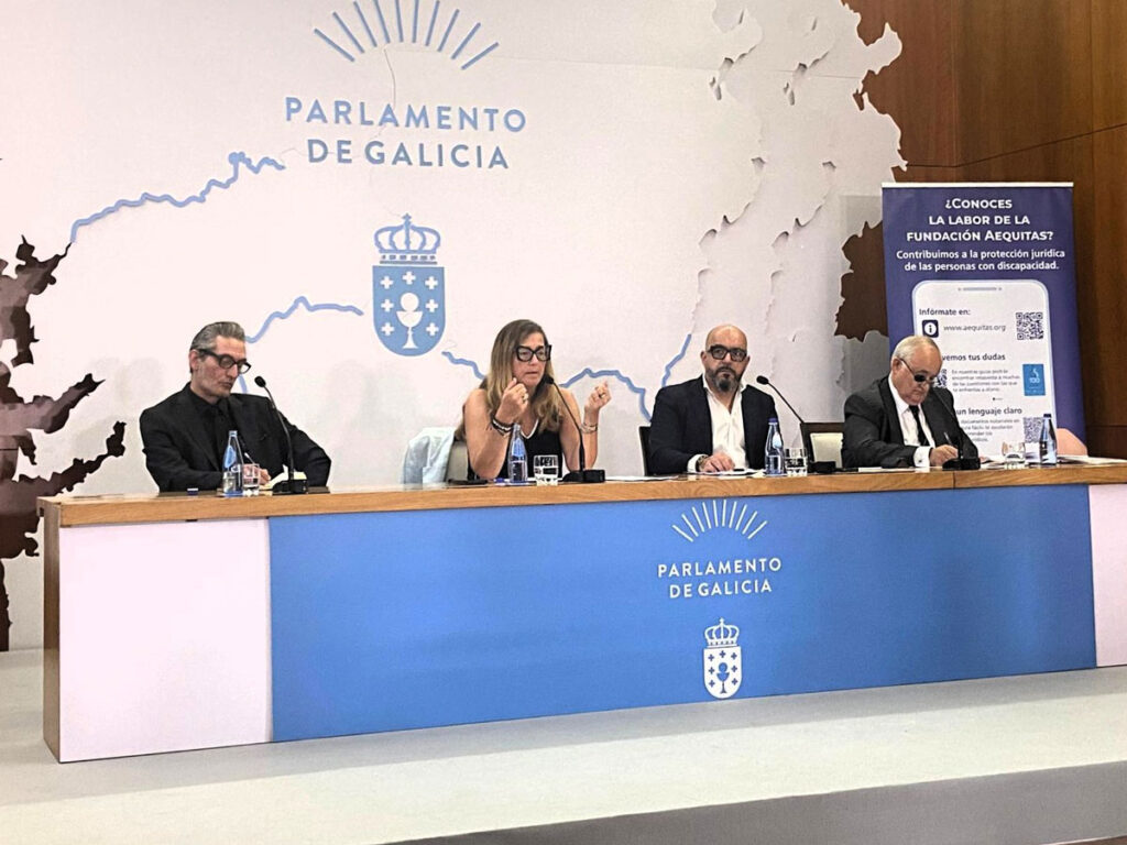 Cuatro personas están sentadas en una mesa alargada con micrófonos, frente a un telón en el que se lee «Parlamento de Galicia». A la derecha se ve un cartel sobre la labor de una fundación. Los ponentes parecen estar inmersos en un debate.