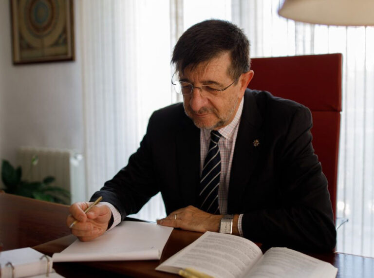 Un hombre con traje y gafas está sentado ante un escritorio, escribiendo en una hoja de papel con un libro abierto delante de él. Parece concentrado, con la luz que entra por una ventana a sus espaldas. Medidas no judiciales y sus efectos.