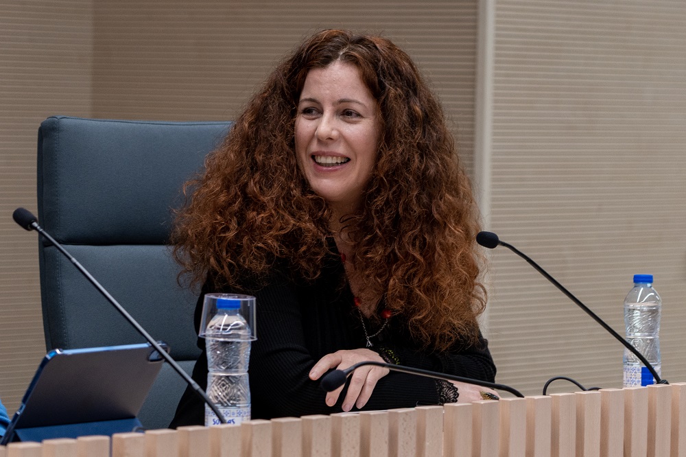 Una mujer con el pelo largo, rizado y de color castaño rojizo sonríe mientras está sentada en una mesa de reuniones junto a unos micrófonos y unas botellas de agua. Jóvenes y salud mental: necesidades de apoyo y retos pendientes en la aplicación de la Ley 8/2021